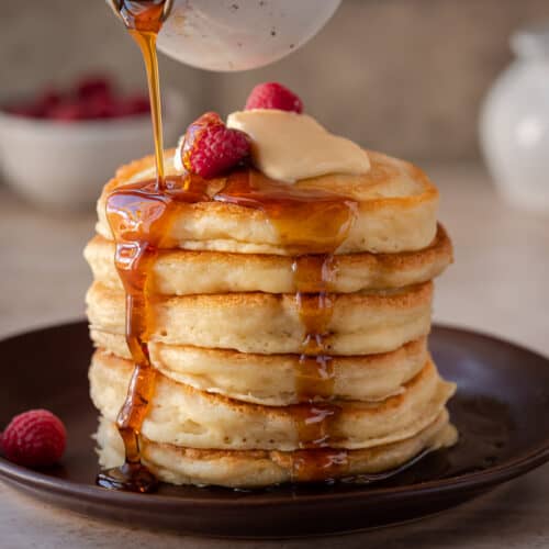 stack of fluffy vegan pancakes on a brown plate with maple syrup being drizzled on top.