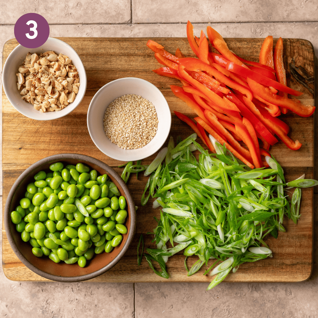 prepped veggies, sesame seeds and peanuts on a wooden cutting board.